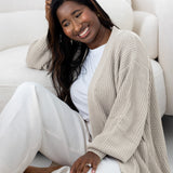 Woman sitting on a white couch wearing a beige cardigan and white pants.