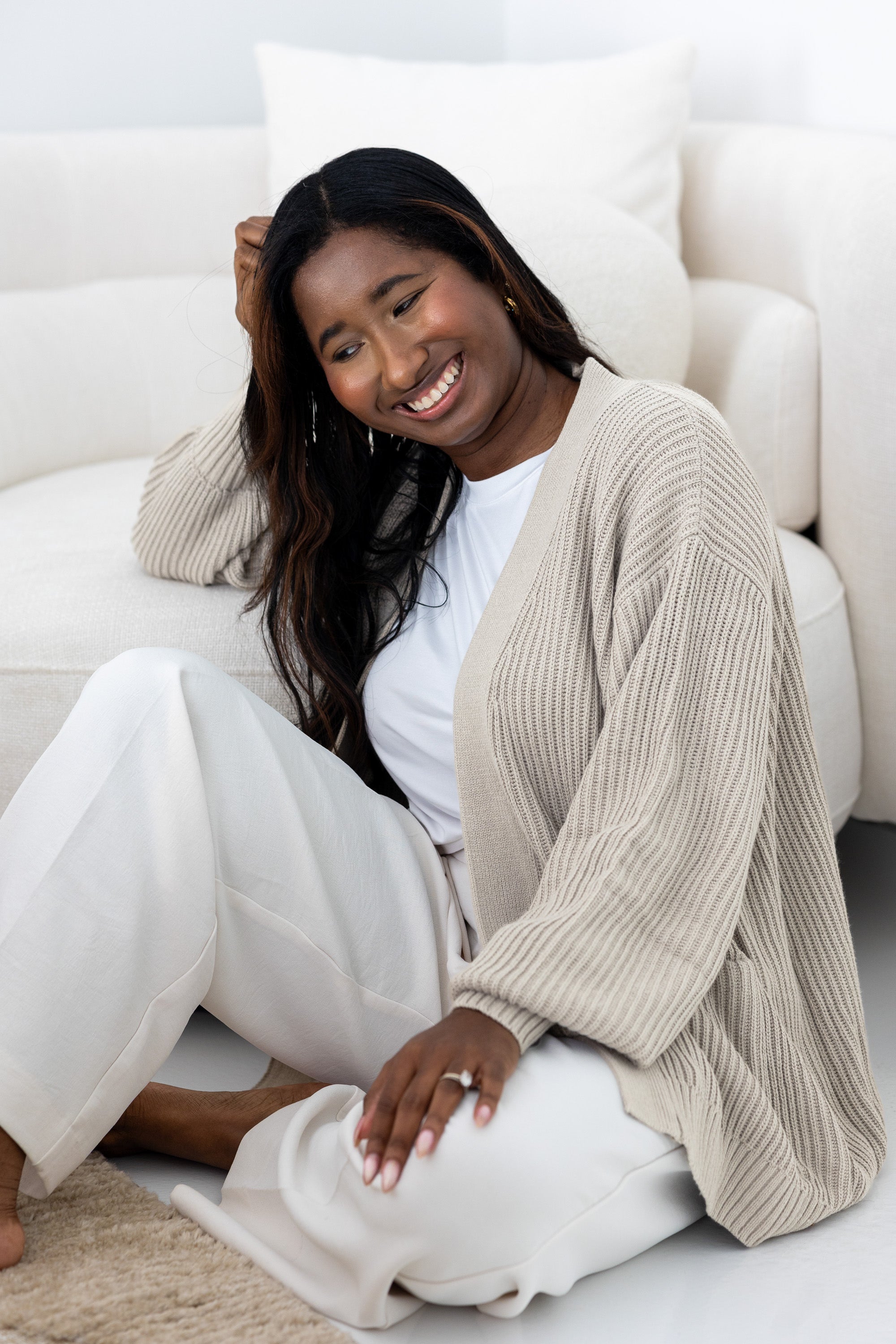 Woman sitting on a white couch wearing a beige cardigan and white pants.