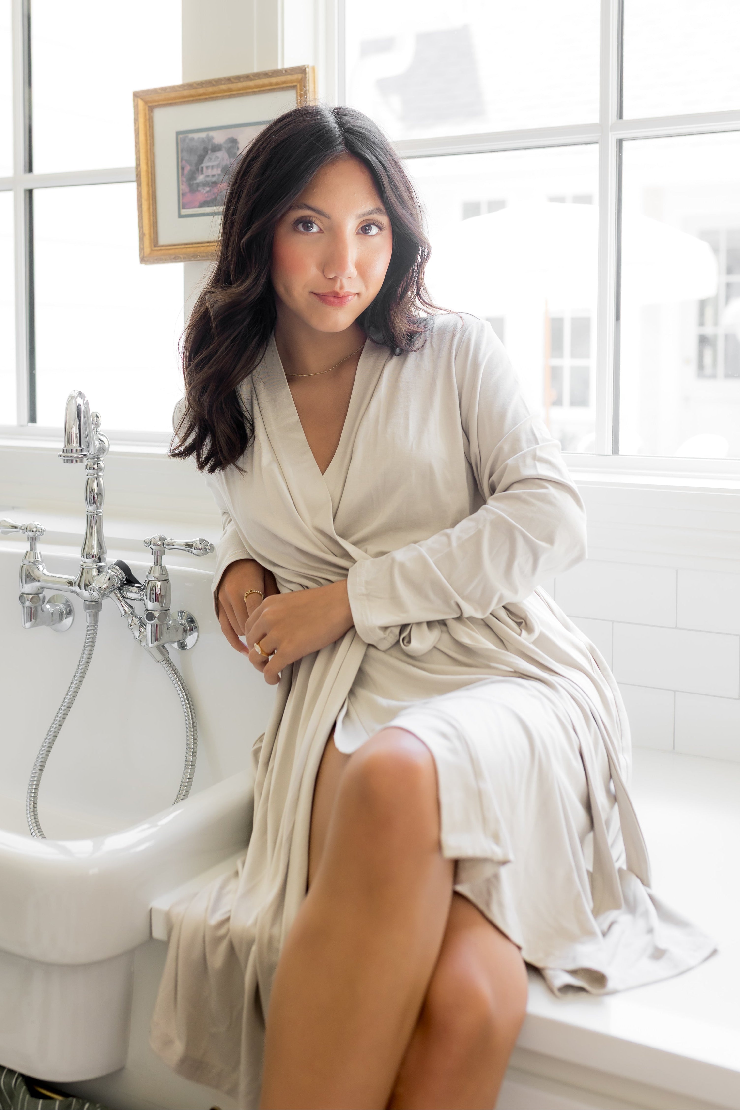 Woman in a off white robe sitting by a window with a sink in the background