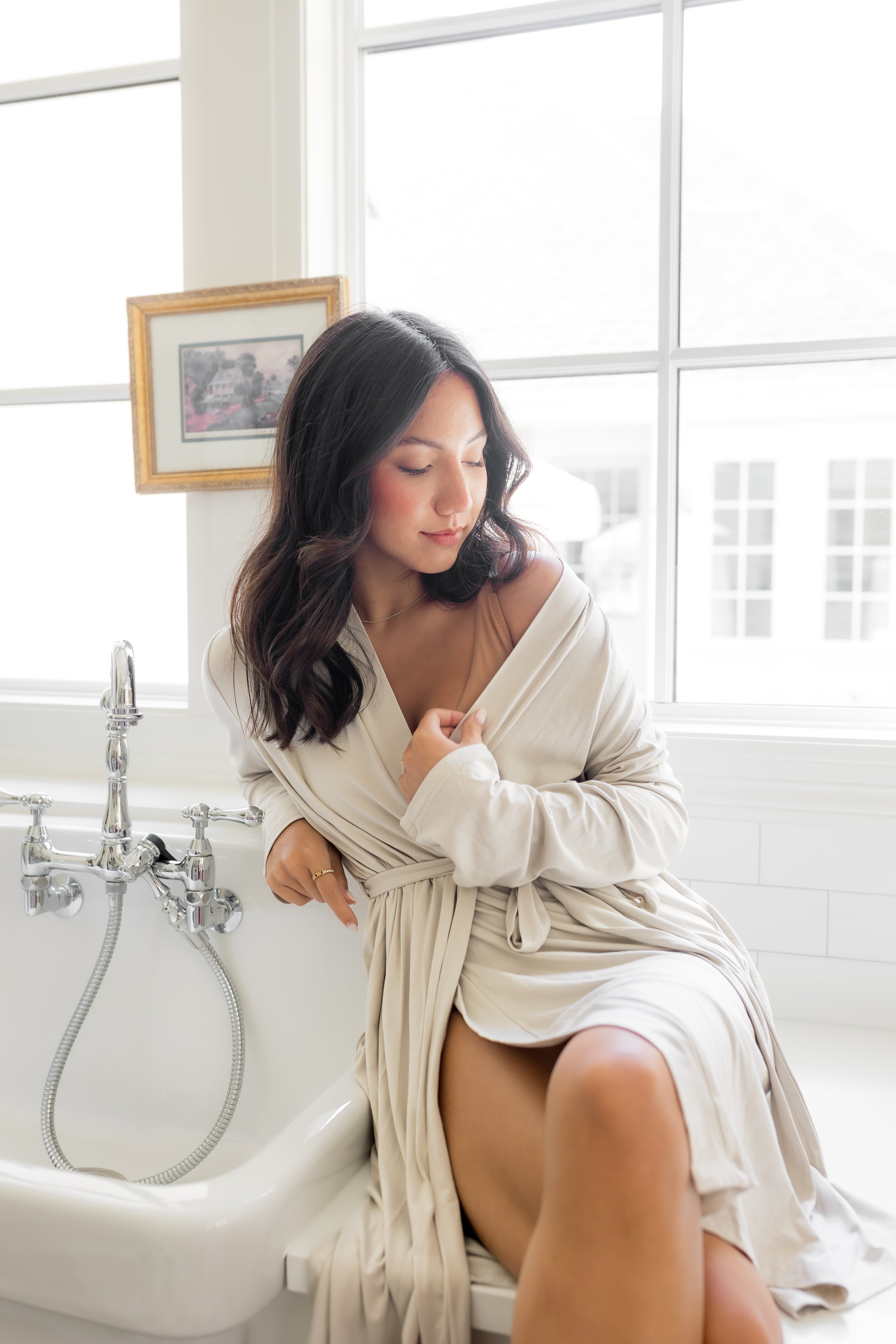 Woman in a off white robe sitting on a bathtub in a bright bathroom.