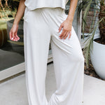 Female model standing on a sidewalk in front of a glass door of a white brick house wearing the Women's Wide Leg Pant in Oat