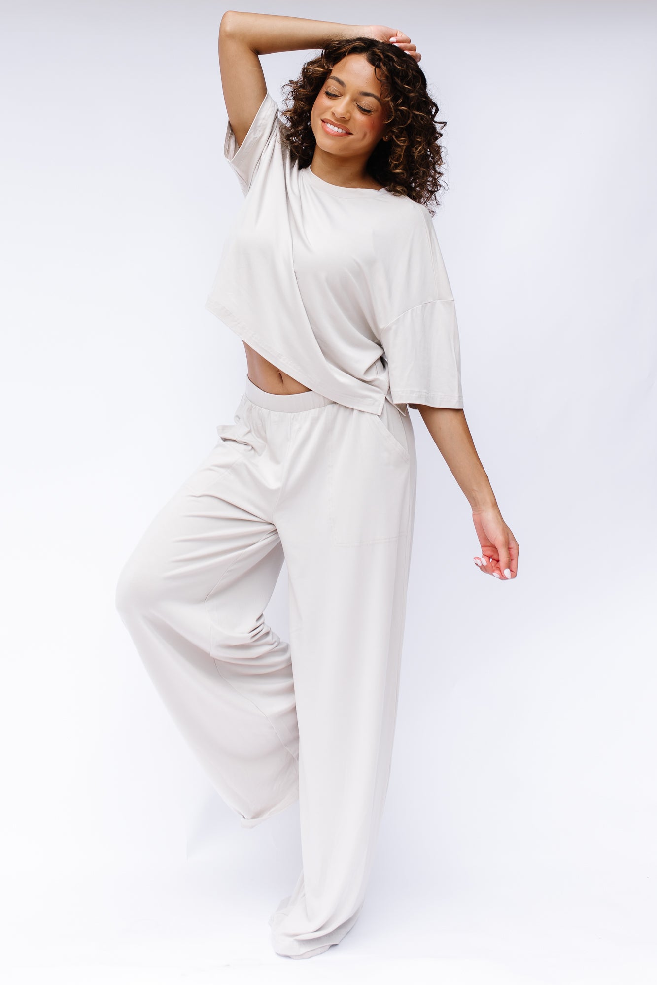 Female model standing with her eyes closed with one leg up and bent and one hand on top of her head wearing the Women's Short Sleeve Tee in Oat and matching wide leg pants