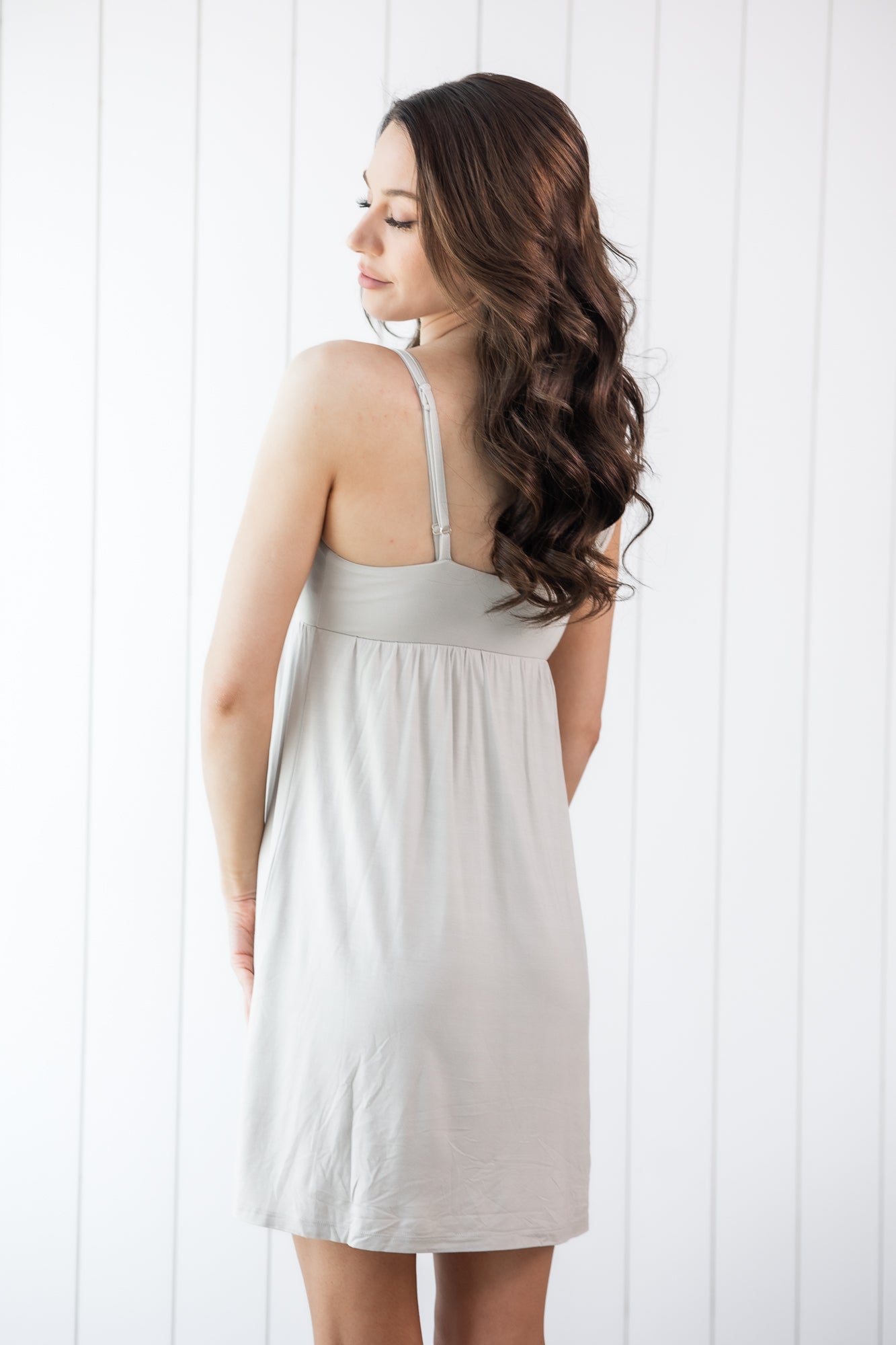 Woman wearing a light gray dress against a white wooden panel background showing the back of the dress