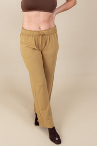 Bottom half of a woman with her leg in front of the other and her hand on her hip wearing sweatpants in ochre and sports bra in espresso