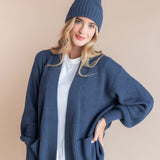 Modal wearing Chunky Knit Adult Ribbed Beanie in oxford over white t-shirt and chunky knit women's cardigan in oxford and blue jeans on a beige background