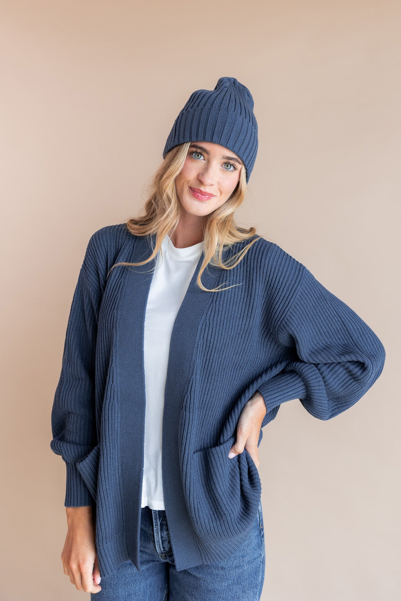 Modal wearing Chunky Knit Adult Ribbed Beanie in oxford over white t-shirt and chunky knit women's cardigan in oxford and blue jeans on a beige background