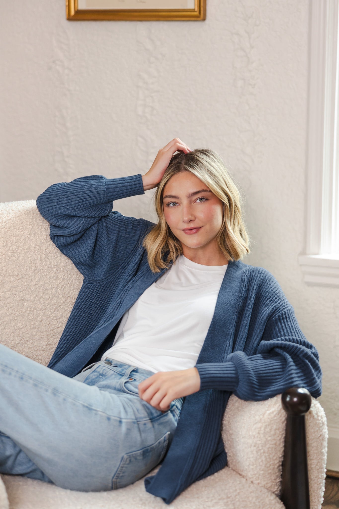 Woman sitting on white sofa wearing an oversized cardigan in oxford over white t-shirt and blue jeans
