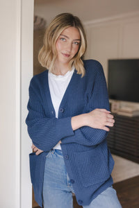 Modal standing against a wall wearing Chunky Knit Women's Waffle Cardigan in Oxford over white t-shirt and blue jeans