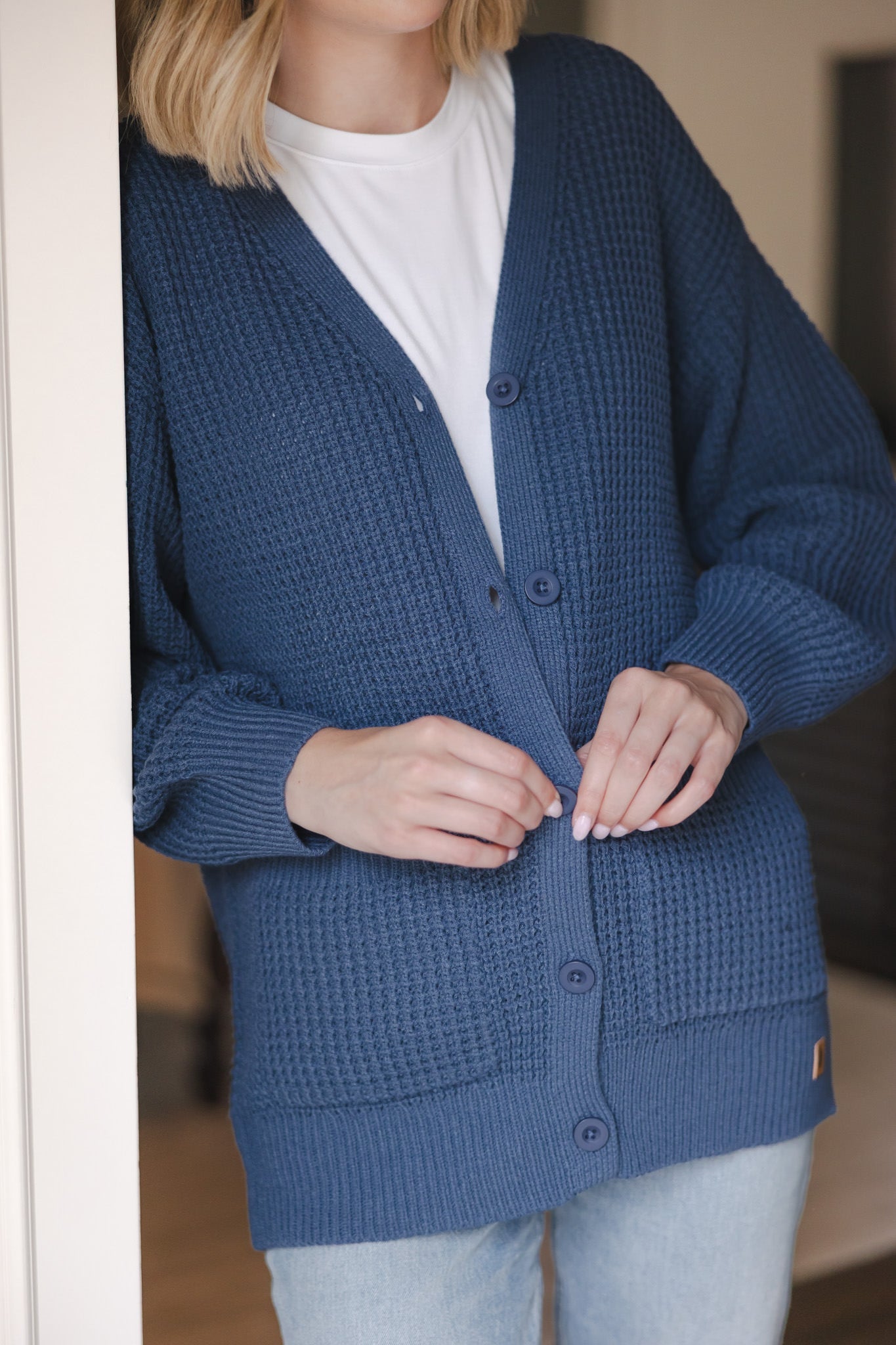Close up of Modal standing against a wall wearing Chunky Knit Women's Waffle Cardigan in Oxford with buttons over white t-shirt and blue jeans