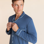 Male model wearing oxford blue button up long sleeve pajamas in a studio setting, adjusting buttons