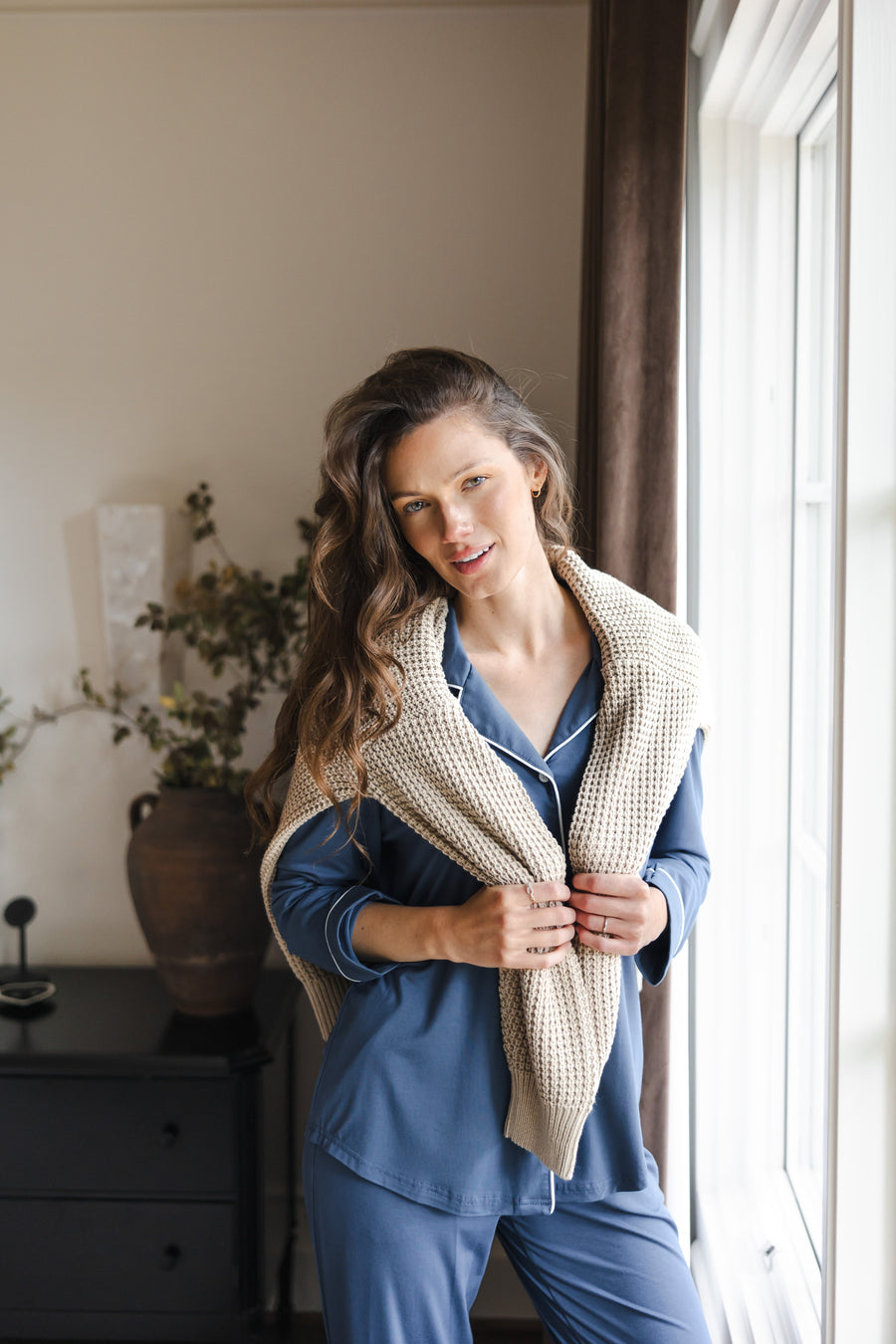 Model smiling in  Long-Sleeved Women's Pajama Set in Oxford with beige sweater over her shoulders standing against a large window
