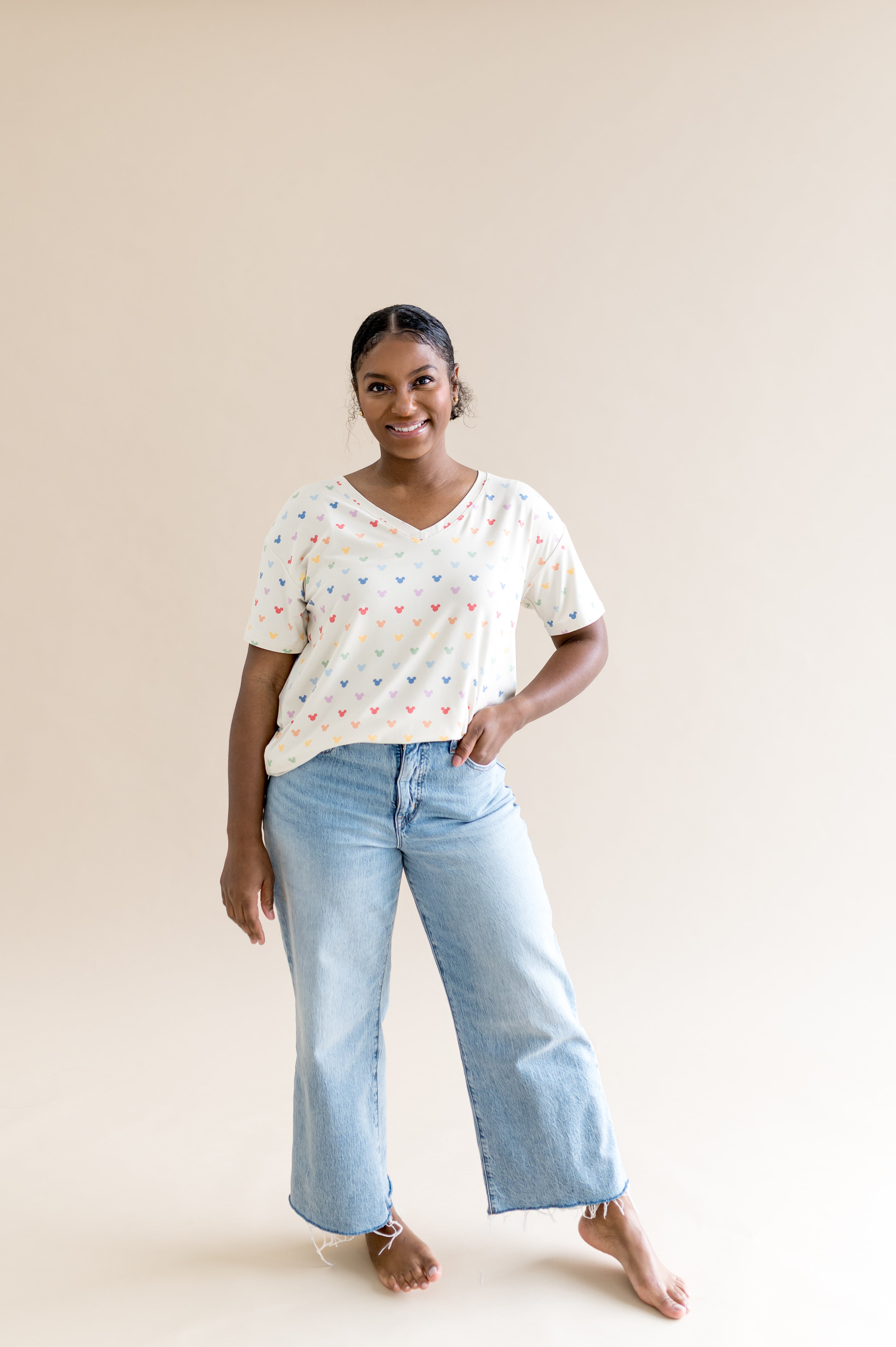 Smiling woman standing in front of a beige background while modeling the Women's Relaxed Fit V-Neck in Pastel Mickey Mouse 