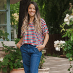 Model standing in front of greenery while wearing the  Women's Relaxed Fit V-Neck in Patriotic Gingham paired with dark denim 