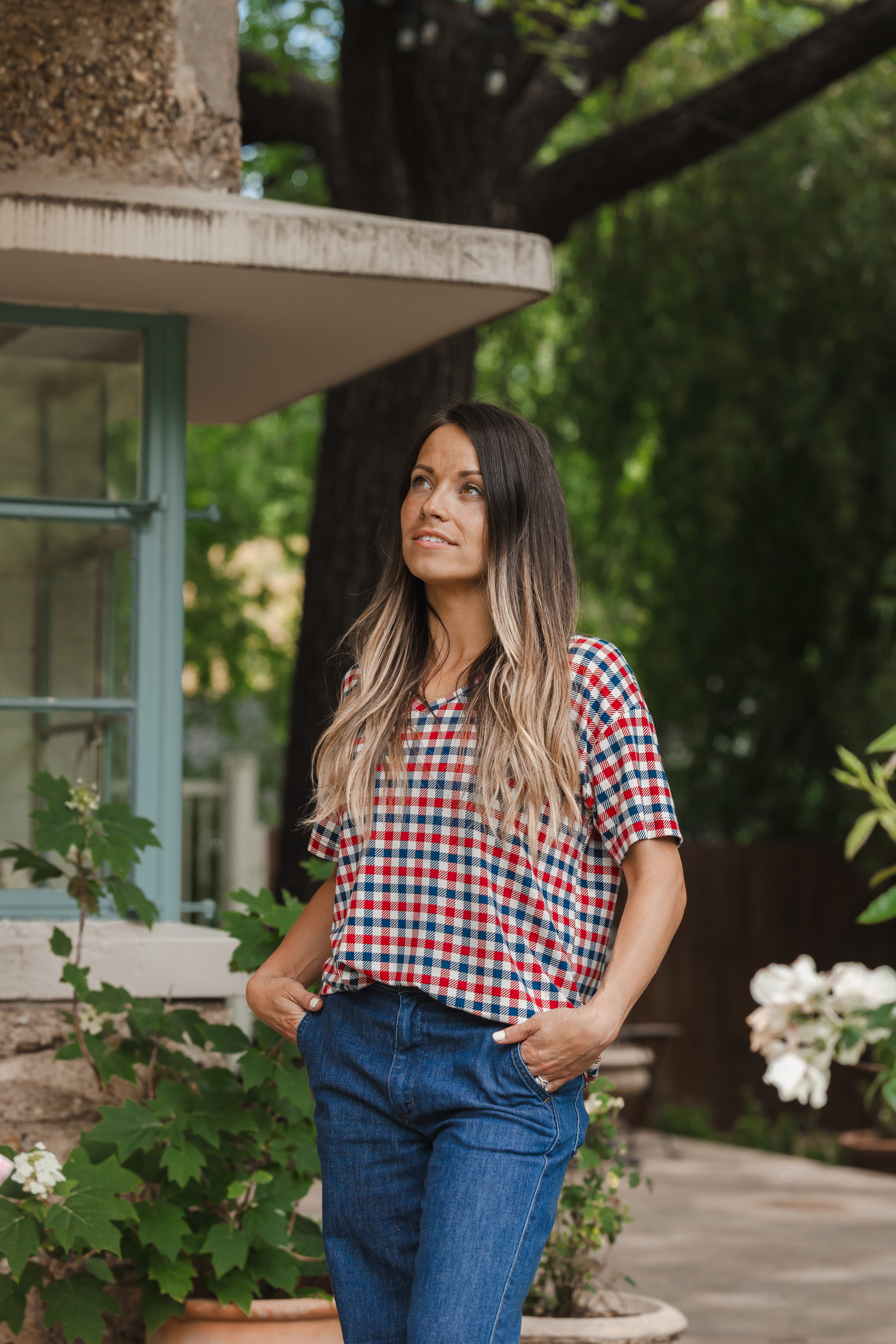 Model standing outside looking upwards while wearing the Women's Relaxed Fit V-Neck in Patriotic Gingham with hands in her jean pockets 