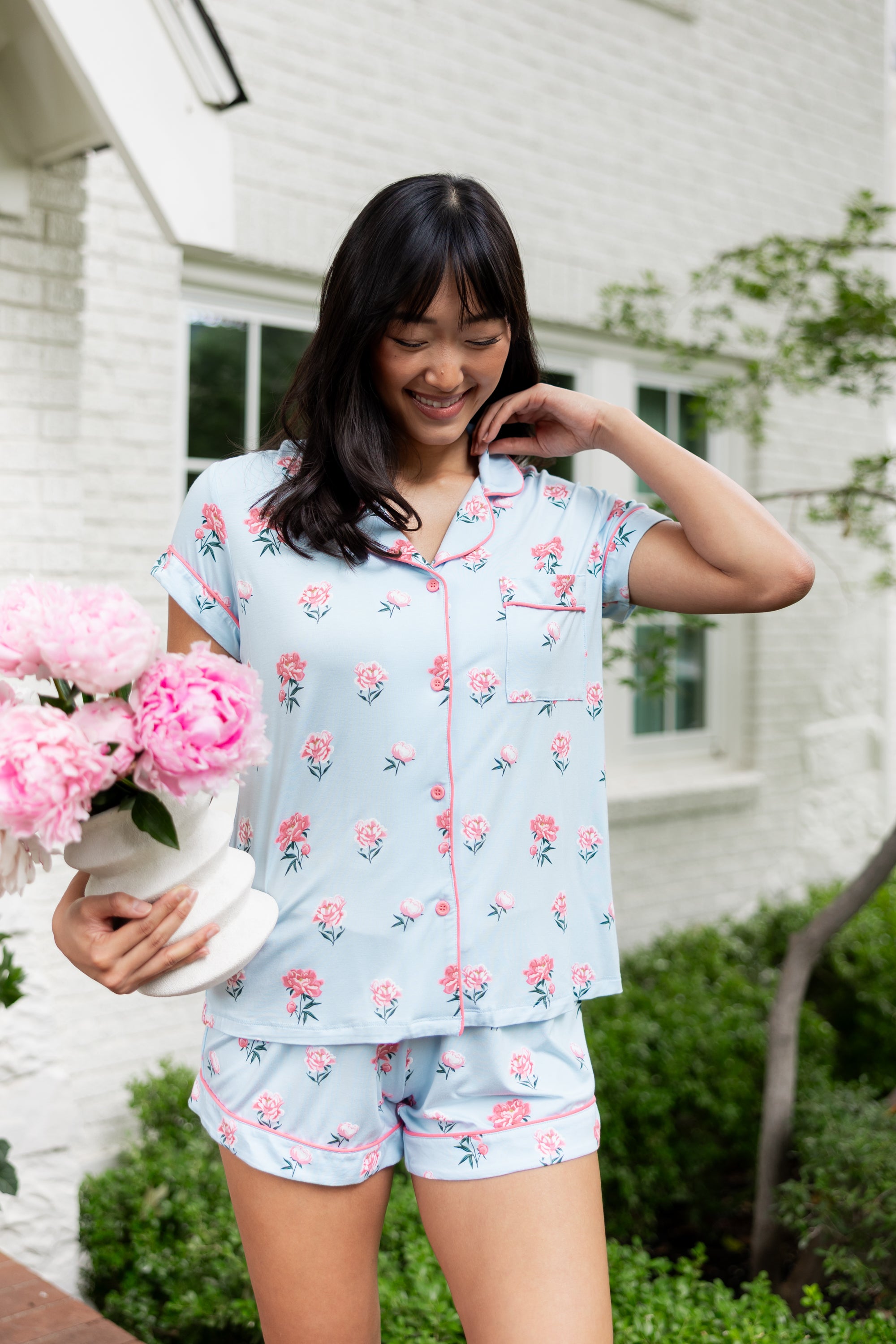 Woman outside smiling down holding a vase of flowers and pushing back her hair in short sleeve pajamas and shorts in Peony on Breeze. ---1615 1