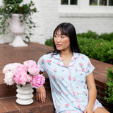 Woman sitting outside on porch steps looking over ger shoulder with a vase of flowers next to her in short sleeve pajama top and shorts in Peony on Breeze print. ---1615 3