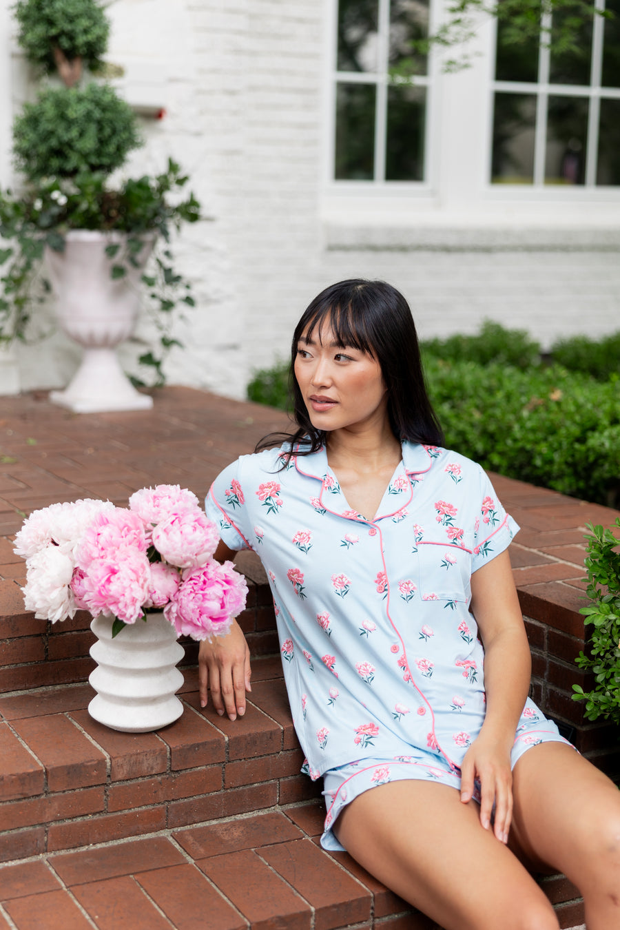 Woman sitting outside on porch steps looking over ger shoulder with a vase of flowers next to her in short sleeve pajama top and shorts in Peony on Breeze print.