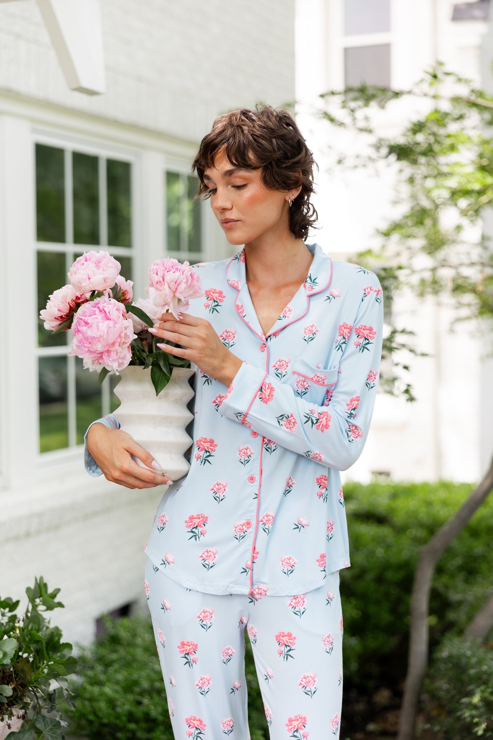 A woman wearing a light blue pajama set with a pink and green floral print stands outdoors in front of a white house, holding a white vase with pink flowers.
