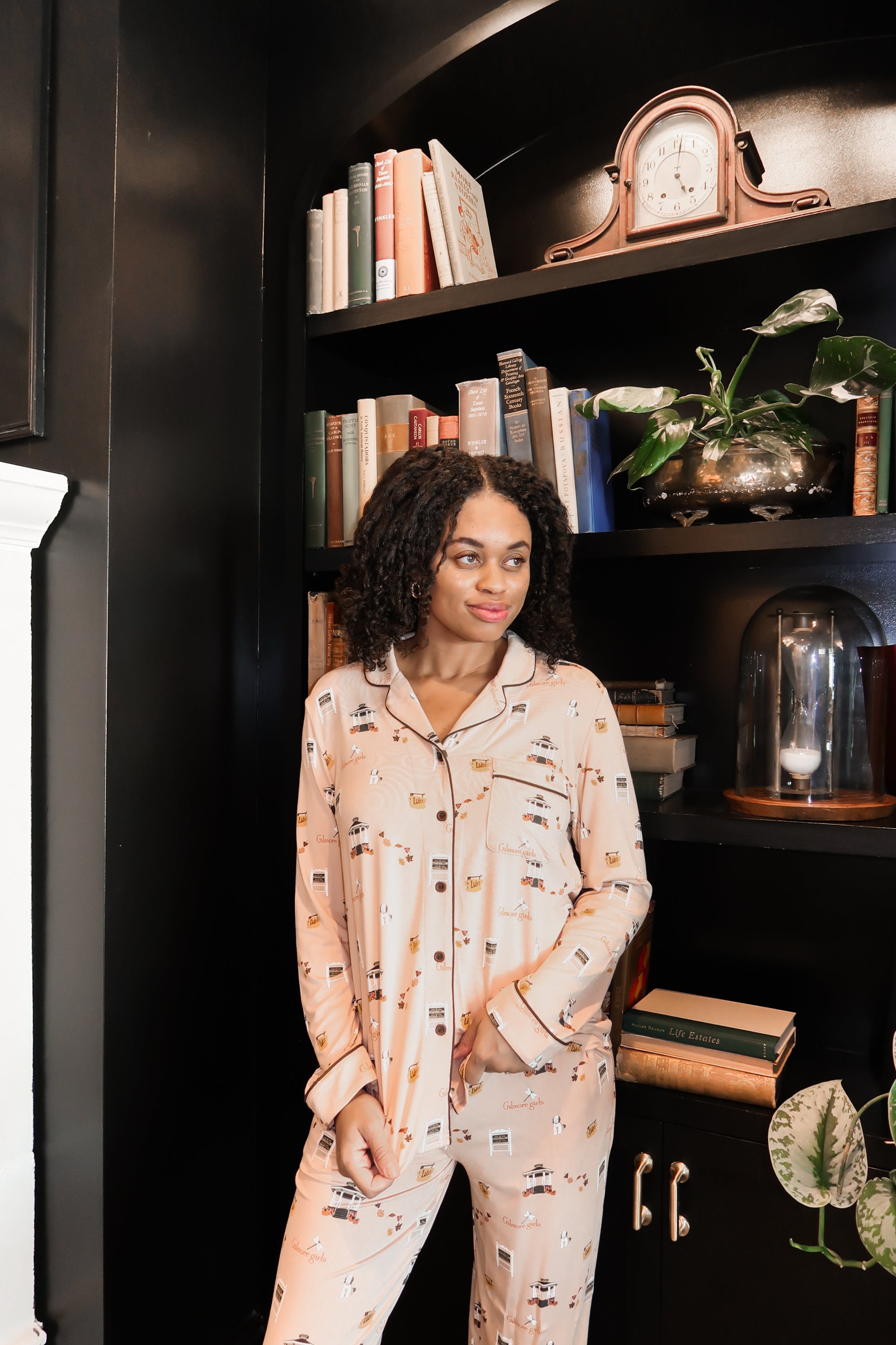 Woman in gilmore girl printed pajamas standing in front of a bookshelf with decorative items.