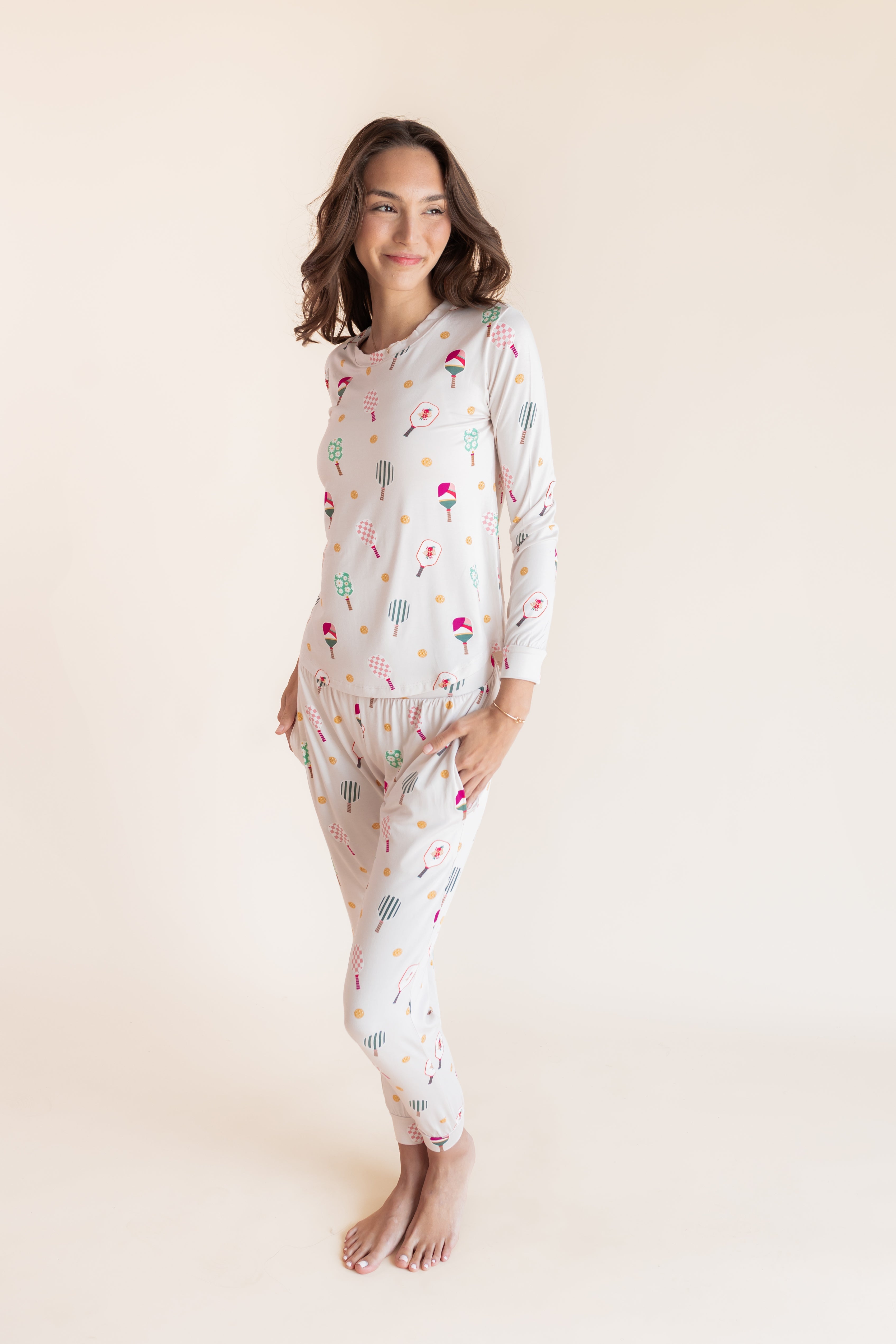A woman modeling a two-piece long-sleeve pajama set with a colorful pickleball print on a light cream background, set against a solid warm beige studio backdrop.
