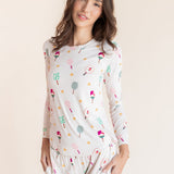 A woman modeling a two-piece pajama set featuring a long-sleeve top and matching pants with a colorful pickleball pattern on a white background, set against a solid light beige backdrop.