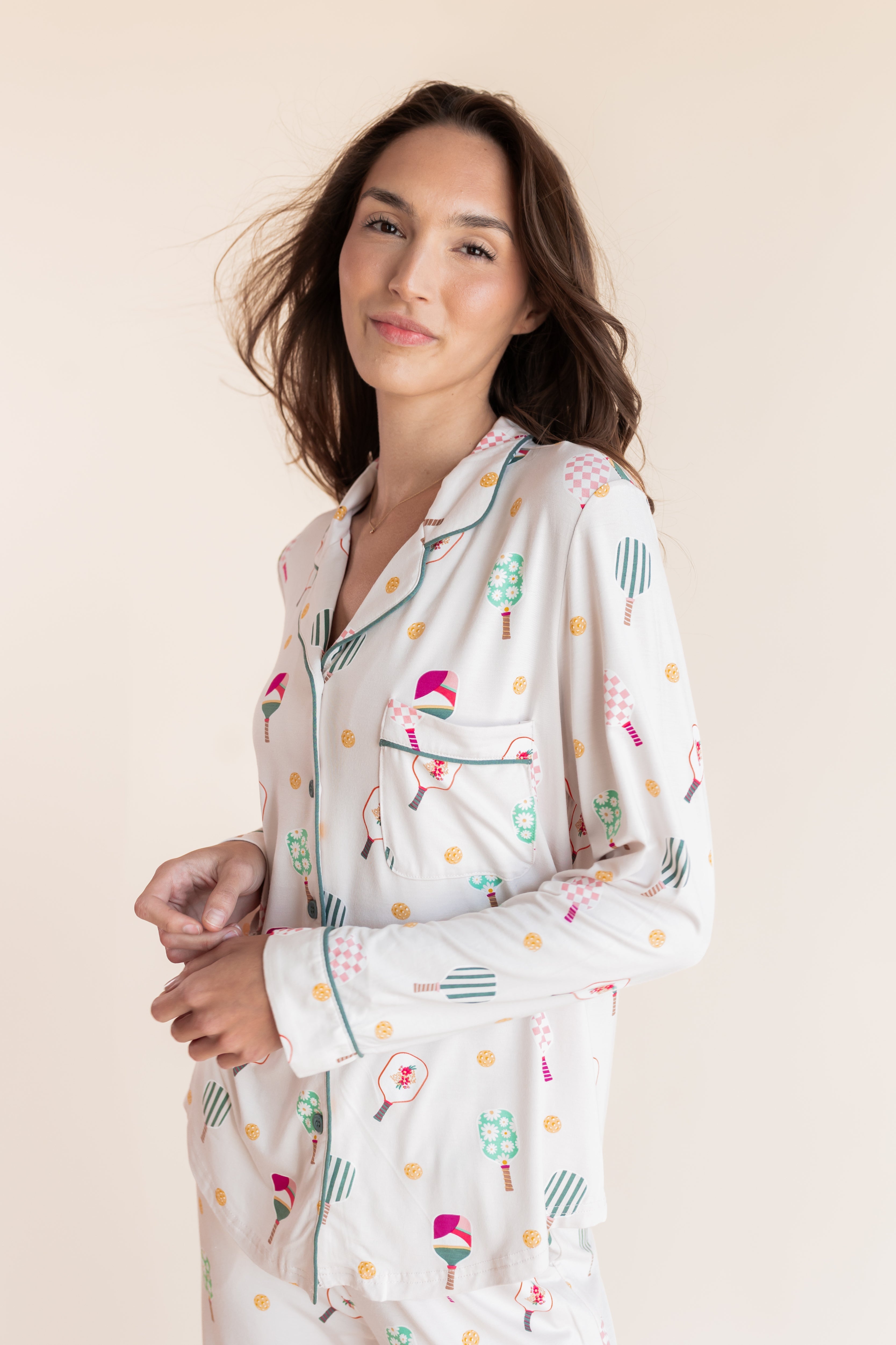 A woman wearing a long-sleeve pajama shirt with a colorful pickleball pattern on a cream background, posed against a solid light beige backdrop.
---PJ 4