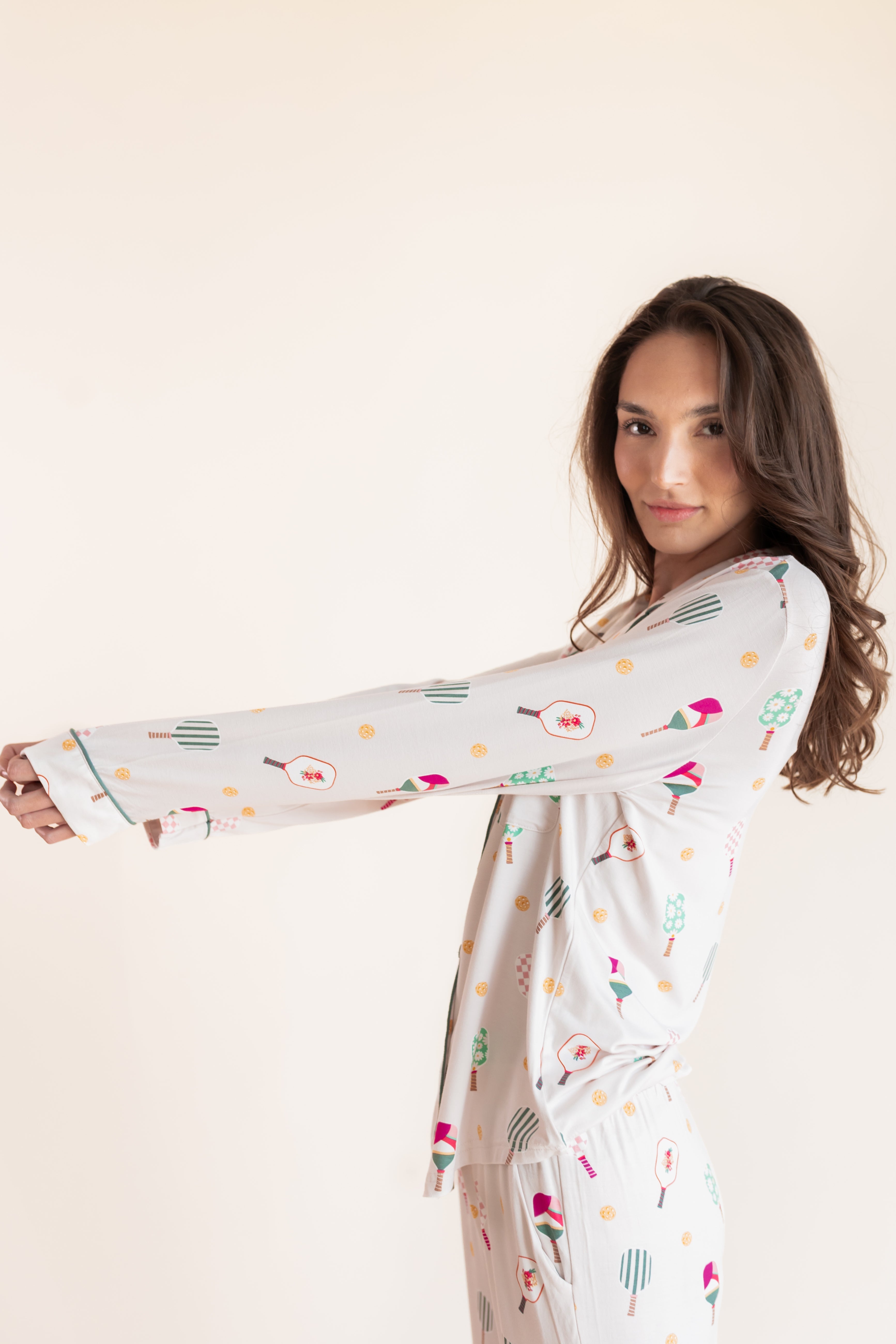 A woman modeling a long-sleeve white pajama set with a colorful pickleball  print, posed against a solid light beige background.
---1605 1