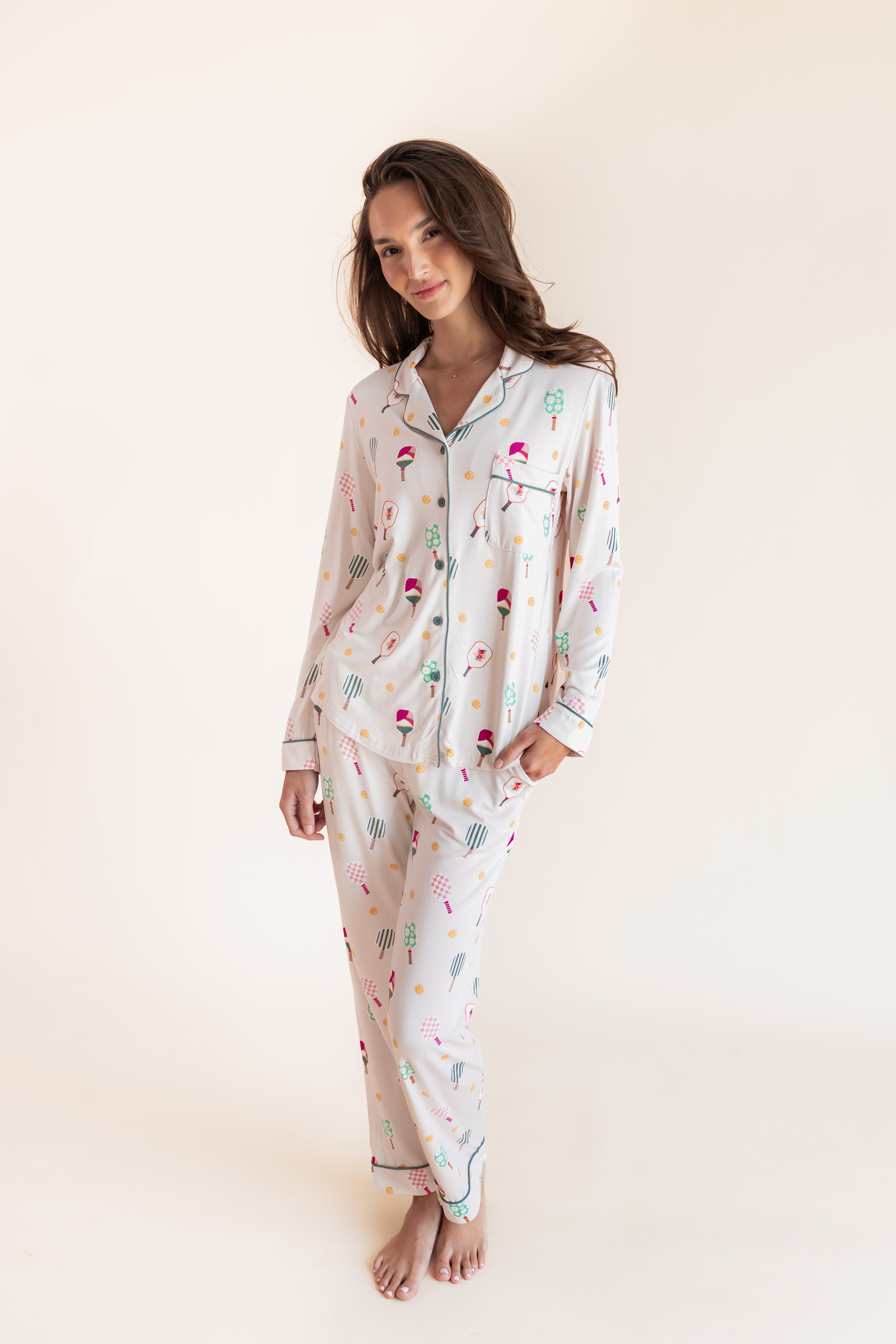 A woman modeling a matching long-sleeve pajama set with a white base and a colorful pickleball print against a plain cream studio background.

