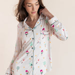 A woman wearing a long-sleeve button-front pajama top and matching pajama pants with a whimsical multicolor print of pickleball rackets on a cream background, set against a solid light beige studio backdrop.
---PJ 2