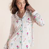 A woman wearing a long-sleeve button-front pajama top and matching pajama pants with a whimsical multicolor print of pickleball rackets on a cream background, set against a solid light beige studio backdrop.
---PJ 2