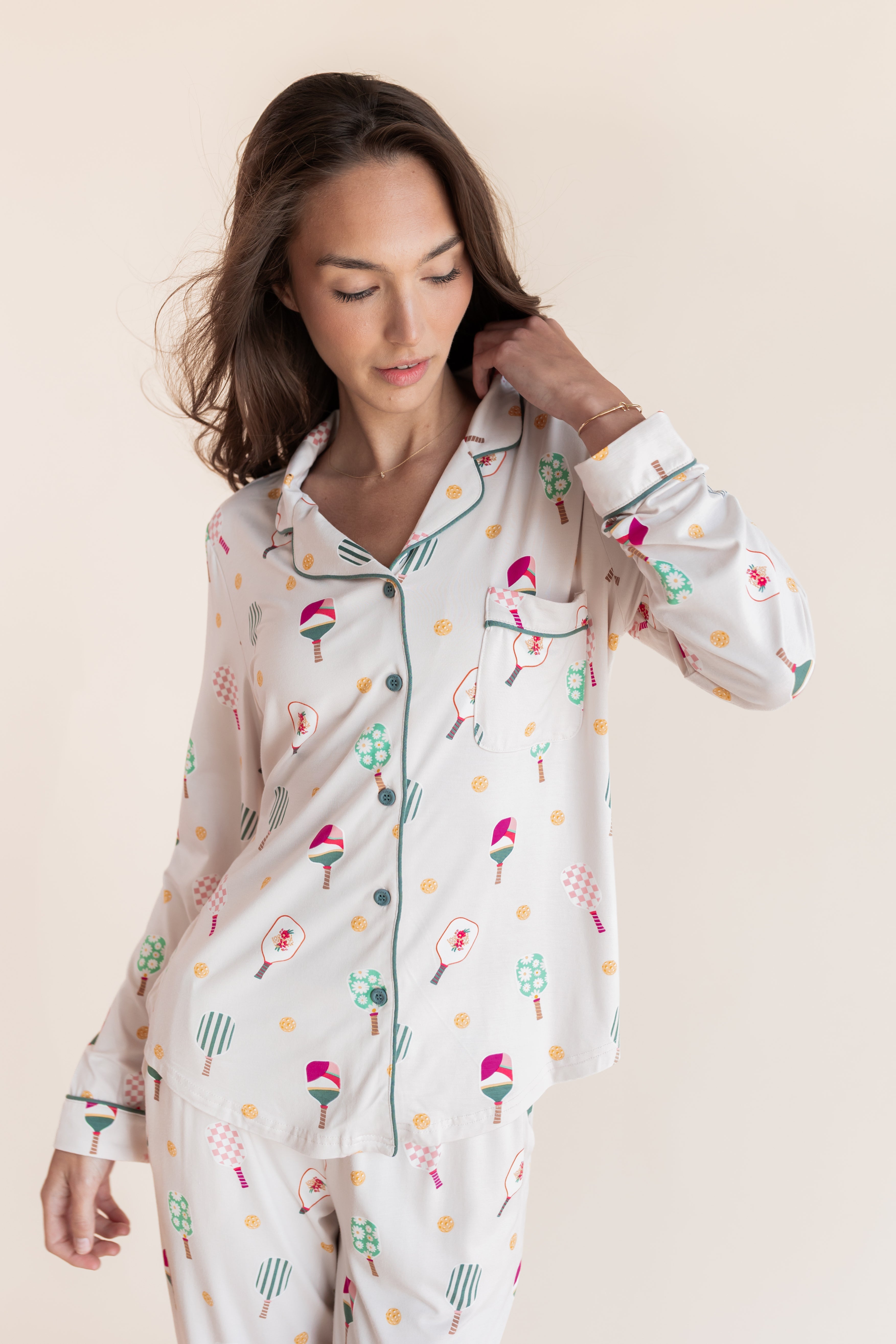 A woman wearing a long-sleeve button-front pajama top and matching pajama pants with a whimsical multicolor print of pickleball rackets on a cream background, set against a solid light beige studio backdrop.
---PJ 2