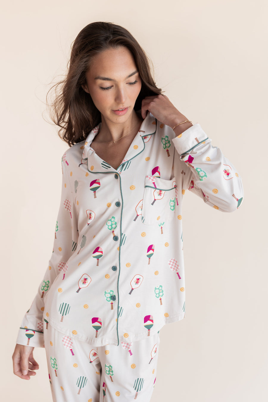 A woman wearing a long-sleeve button-front pajama top and matching pajama pants with a whimsical multicolor print of pickleball rackets on a cream background, set against a solid light beige studio backdrop.