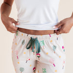 A person wearing cream-colored lounge pants with a colorful pickleball print and a teal drawstring, paired with a white tank top, shown from the chest down against a light beige background.