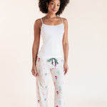 A woman modeling a two-piece pajama set consisting of a white camisole top and white pants with a colorful pickleball  print, set against a solid cream background.