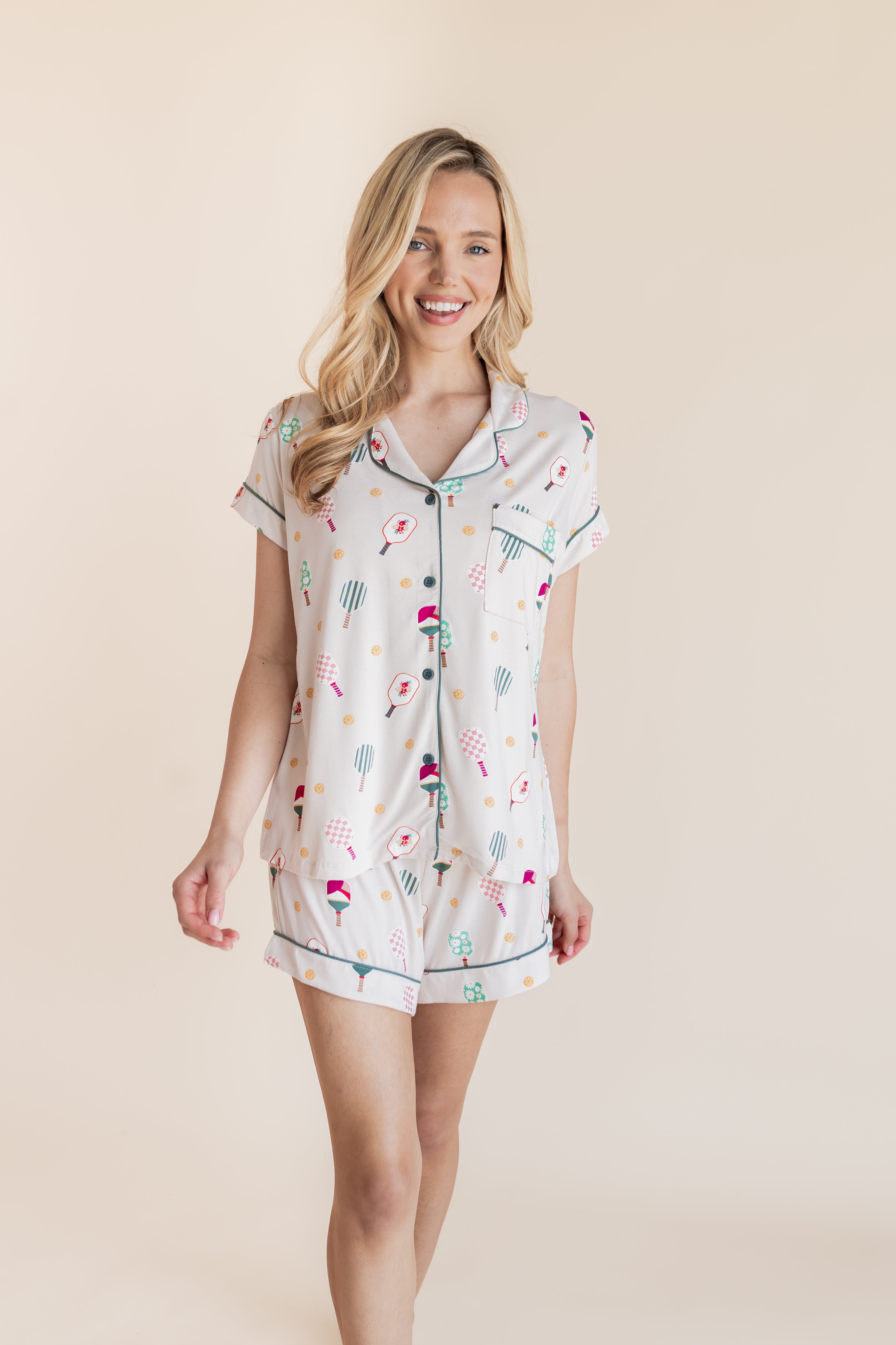 A woman modeling a short-sleeve pajama shirt and matching shorts set with a multicolor pickleball print on a warm off-white background, set against a solid beige studio backdrop.
