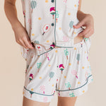 A person wearing a matching short-sleeve button-front pajama top and shorts set in a light cream color with a playful multicolor print of pickleball rackets
---1615 3