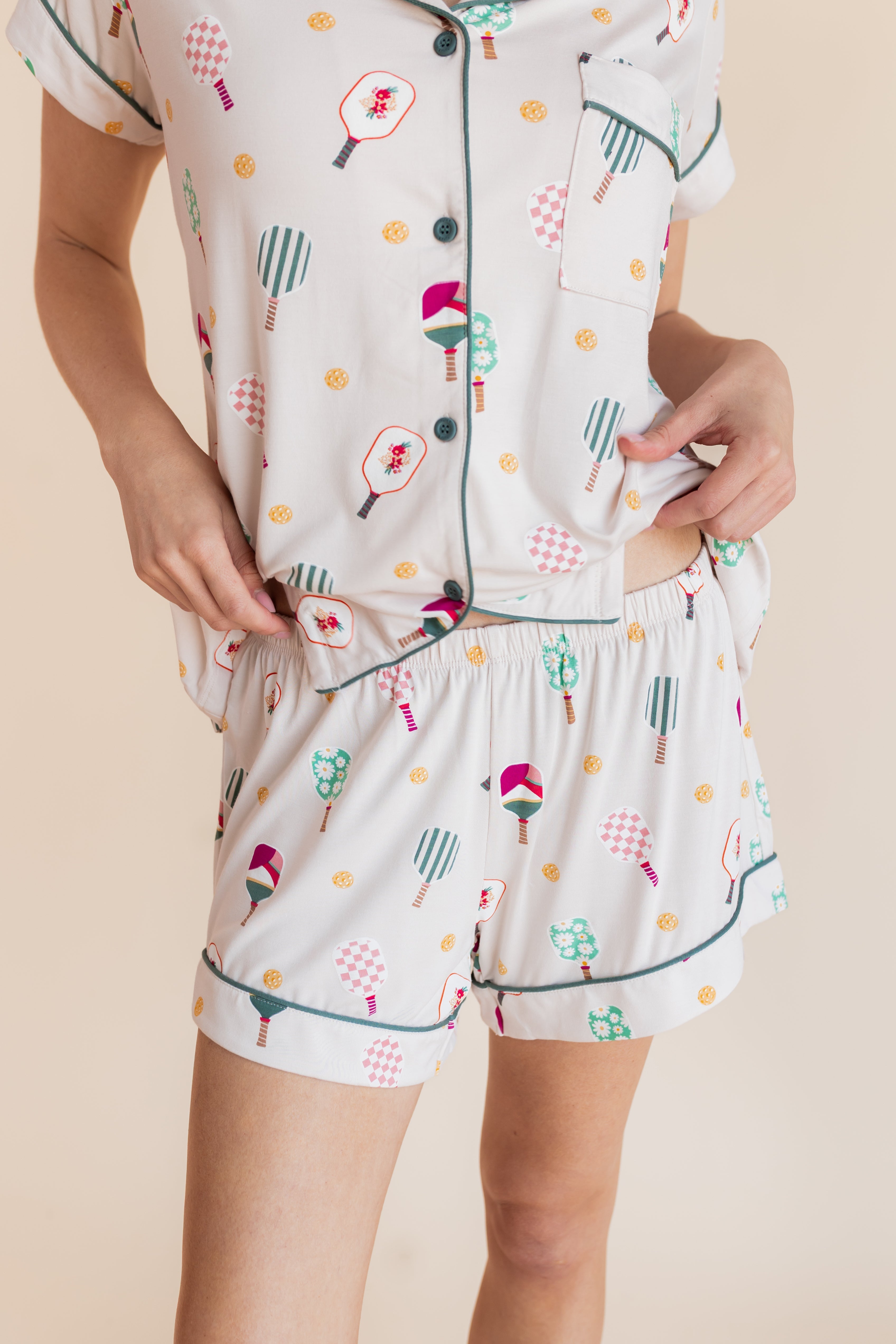 A person wearing a matching short-sleeve button-front pajama top and shorts set in a light cream color with a playful multicolor print of pickleball rackets
---1615 3