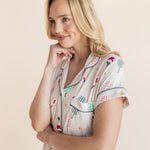 A blonde woman wearing a short-sleeve pajama top with a colorful pickleball-themed print, posing against a solid light beige background.