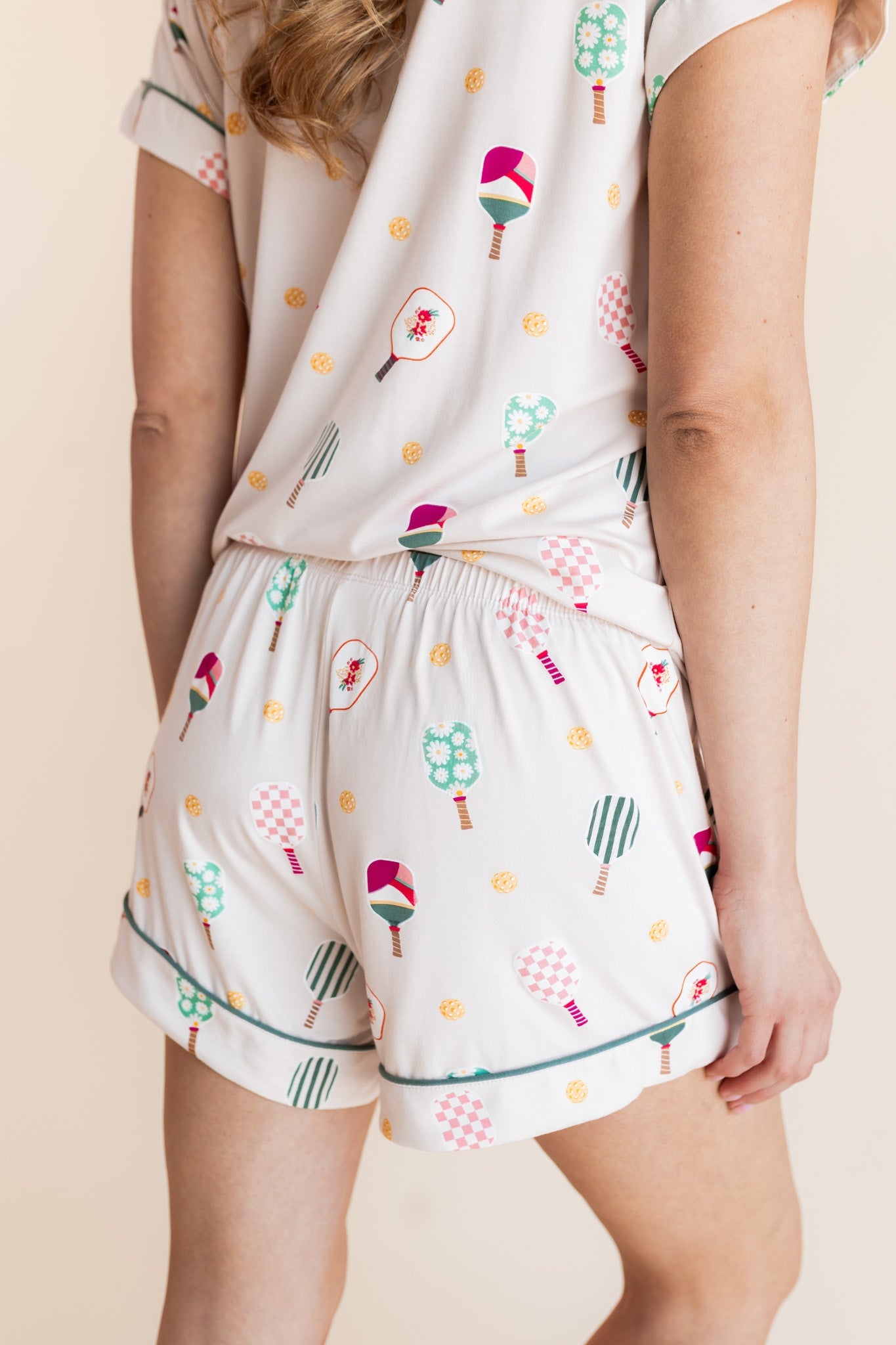 A person wearing a matching two-piece pajama set with a cream background and a colorful pickleball pattern, shown from the back against a plain beige studio background.