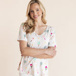 A smiling blonde woman wearing a short-sleeve V-neck top and white scrub pants, featuring a pickleball pattern on a cream background, posed against a solid light beige studio backdrop.
