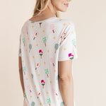 A woman shown from the back wearing a white short-sleeve t-shirt with a colorful pickleball print paired with white pants, against a solid beige background.