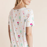 A woman shown from the back wearing a white short-sleeve t-shirt with a colorful pickleball print paired with white pants, against a solid beige background.