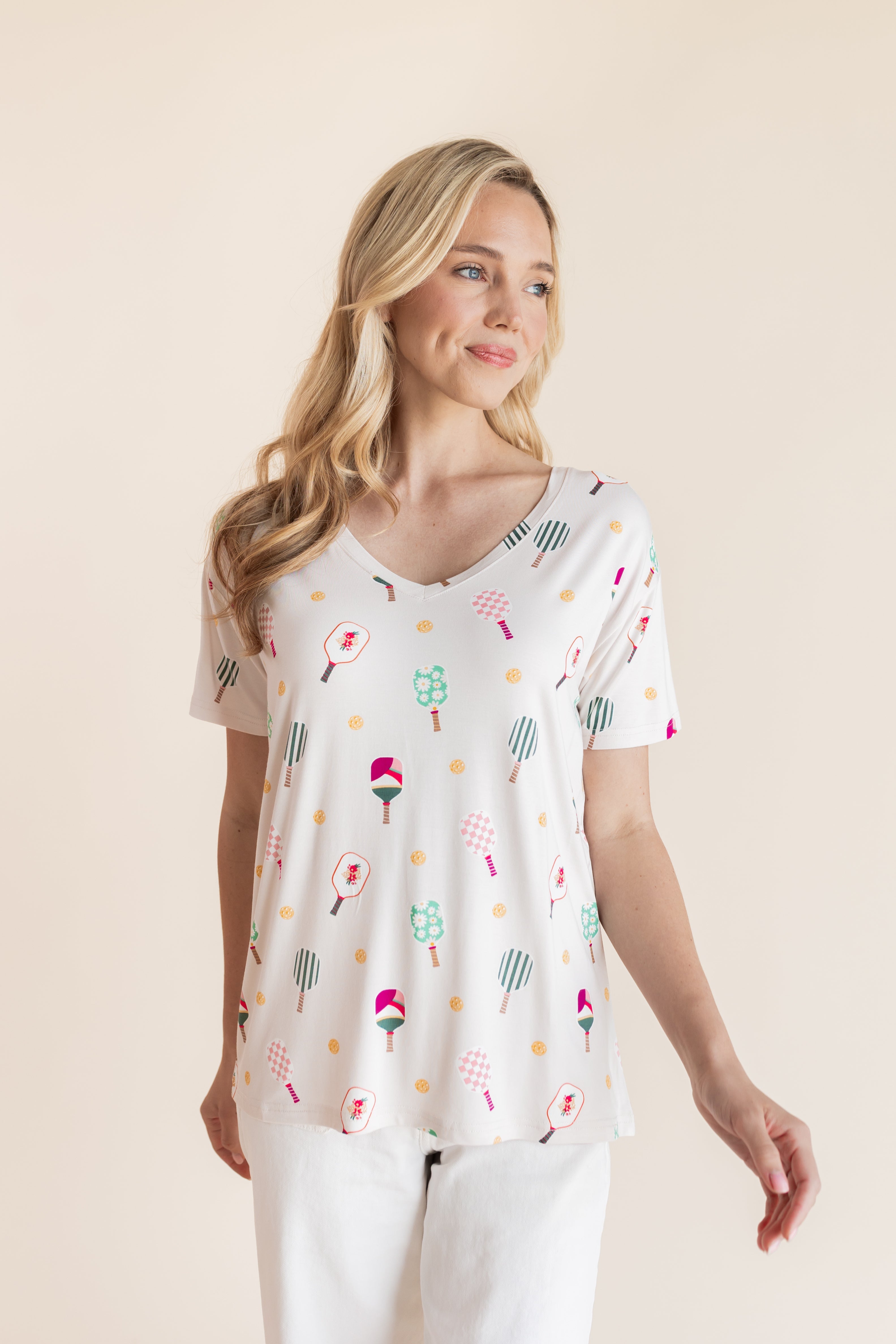 A woman modeling a white short-sleeve V-neck top with a colorful pickleball print, paired with white pants, against a solid beige background.
::: Model is 5'9" wearing size XS ///