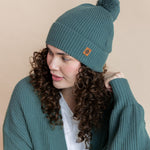 Modal wearing Chunky Knit Adult Pom in Pine over chunky knit women's cardigan in pine  and white t-shirt on a beige background