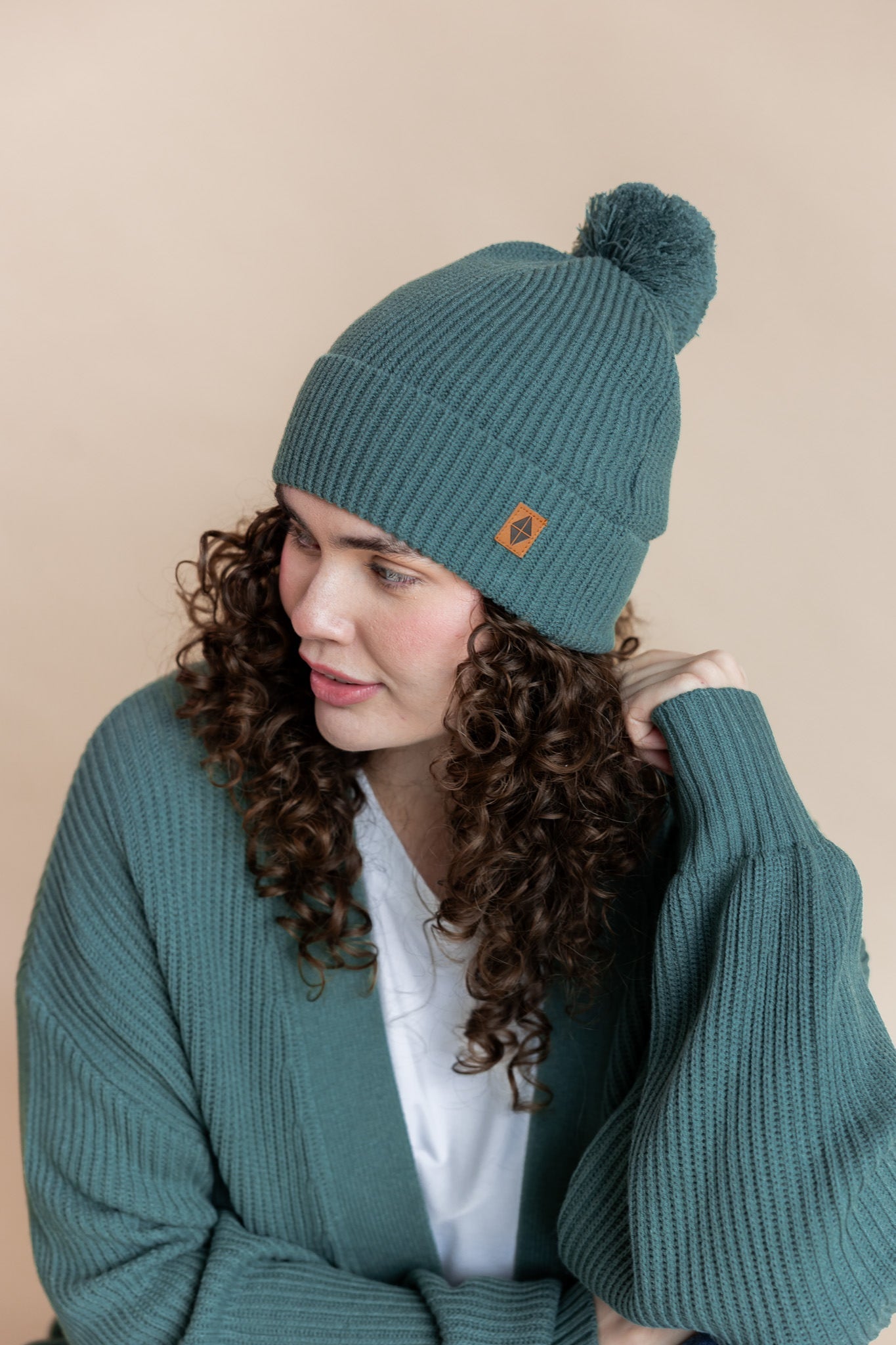 Modal wearing Chunky Knit Adult Pom in Pine over chunky knit women's cardigan in pine  and white t-shirt on a beige background