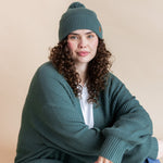 Modal wearing Chunky Knit Adult Pom in Pine over chunky knit women's cardigan in pine, blue jeans and white t-shirt on a beige background