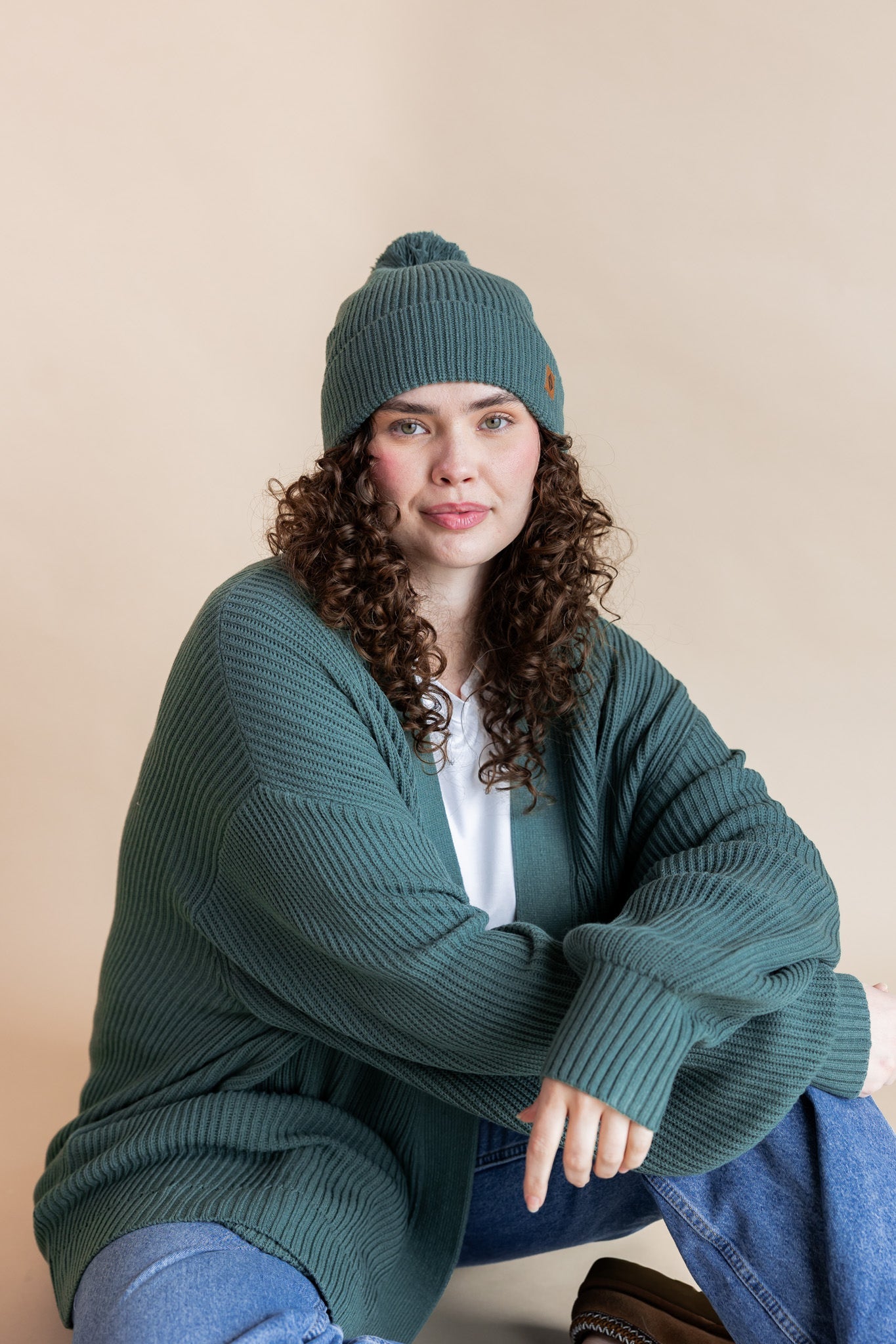Modal wearing Chunky Knit Adult Pom in Pine over chunky knit women's cardigan in pine, blue jeans and white t-shirt on a beige background