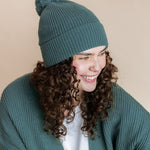 Modal smiling in Chunky Knit Adult Pom in Pine over chunky knit women's cardigan in pine  and white t-shirt on a beige background