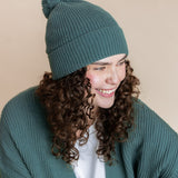 Modal smiling in Chunky Knit Adult Pom in Pine over chunky knit women's cardigan in pine  and white t-shirt on a beige background