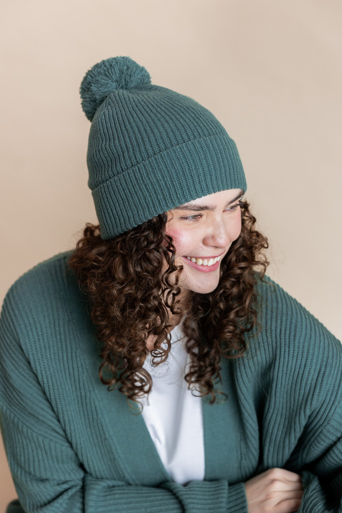 Modal smiling in Chunky Knit Adult Pom in Pine over chunky knit women's cardigan in pine  and white t-shirt on a beige background